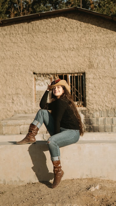 Wearing a brown hat, dressed in a black long sleeved shirt and a blue denim jeans women sit on white chair
